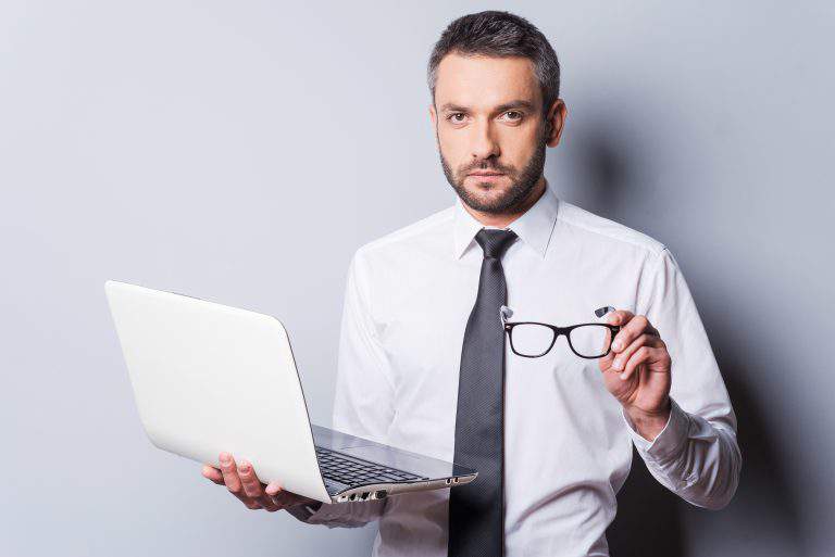 Confident business expert. Confident mature man in shirt and tie holding laptop and eyewear while standing against grey background