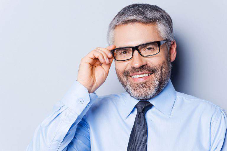 How may I help you? Portrait of confident mature man in shirt and tie adjusting his eyeglasses and smiling while standing against grey background