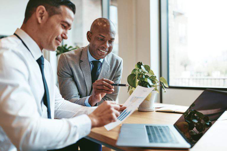 Two smiling diverse businessmen discussing charts and graphs on paper and using a laptop while sitting at a desk in an office