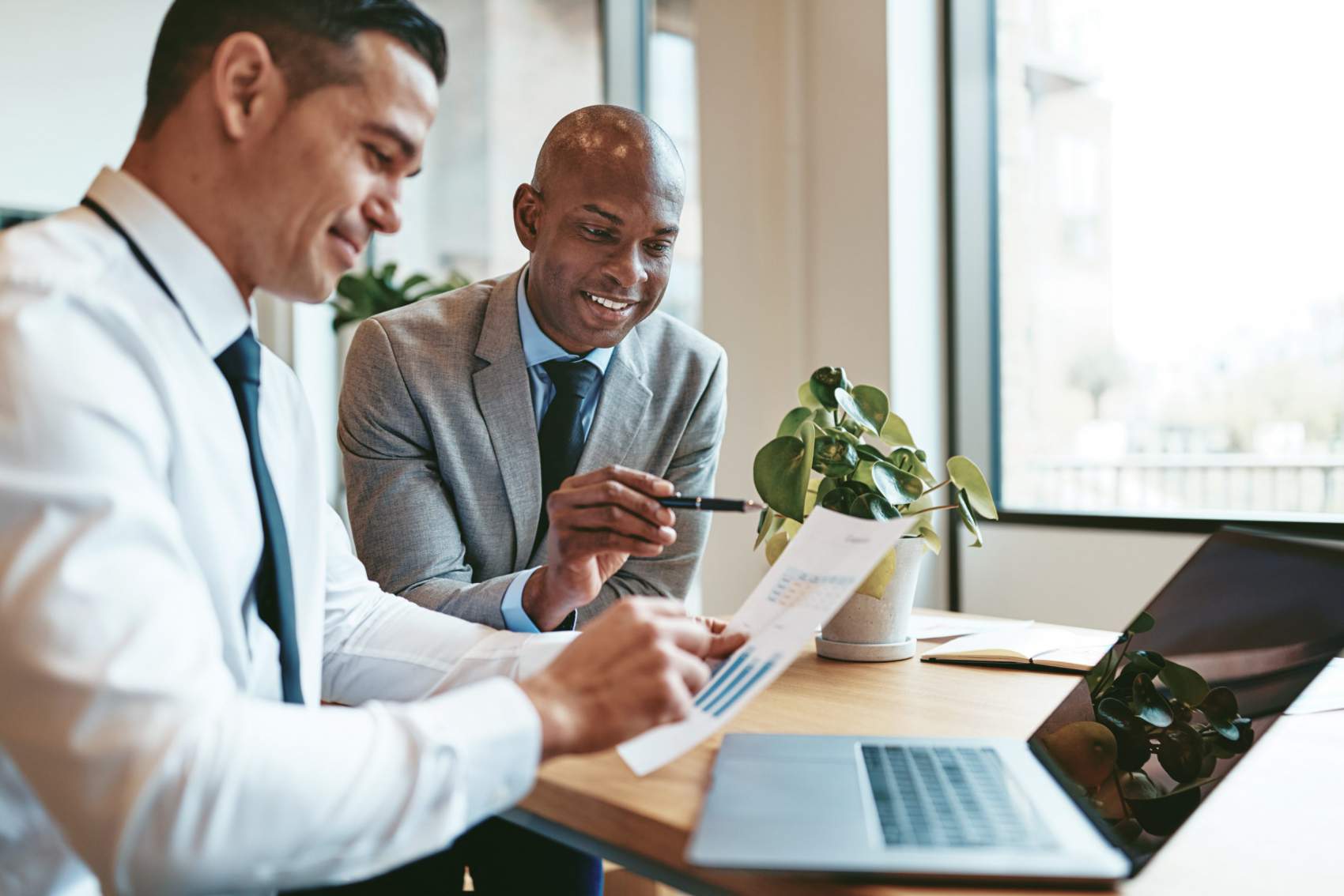 Two smiling diverse businessmen discussing charts and graphs on paper and using a laptop while sitting at a desk in an office