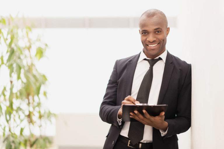 Businessman with note pad. Cheerful young African businessman writing something in his note pad and smiling at camera