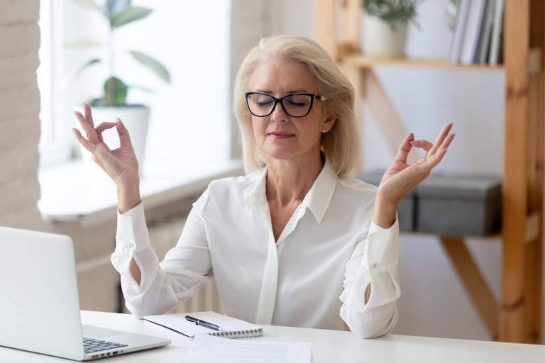 Calm middle-aged businesswoman sit at desk in office relax practice yoga at workplace, peaceful senior female employee meditate control relieve negative emotions, stress free, peace concept